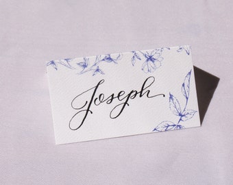 Könnte beinhalten: Weiße Tischkarte mit blauem Blumendesign und dem Namen "Joseph" in schwarzer Kalligraphie.