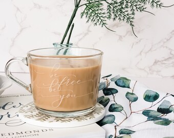 Könnte beinhalten: Eine Glas-Tasse mit weißem Hintergrund und den Worten "be free be you" auf der Seite graviert. Die Tasse ist mit einem hellbraunen Getränk gefüllt und steht auf einem weißen Untersetzer mit grünem Blattdesign. Der Untersetzer liegt auf einem Stapel Bücher mit den Worten "Mastering Hand" und "Edmonds Hill London" auf dem Einband.