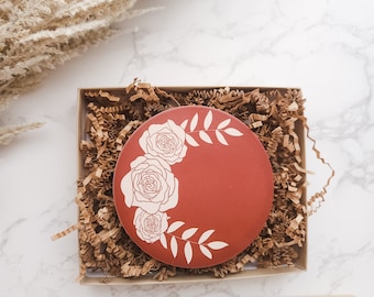 May include: A round terracotta coaster with a white floral design featuring three roses and leaves. The coaster is in a brown paper gift box with brown crinkle paper.