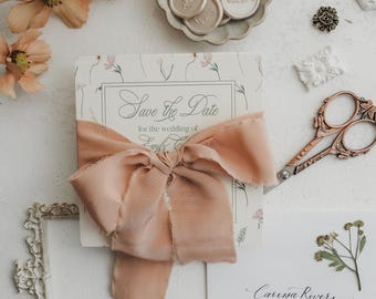 Könnte beinhalten: Eine Hochzeitseinladungssuite mit einer "Save the Date"-Karte mit Pfirsichfarbenem Band, floralen Akzenten und einem passenden Umschlag. Enthält ein goldenes Wachssiegel, dekorative Schere und eine kleine Schale mit Wachssiegeln.