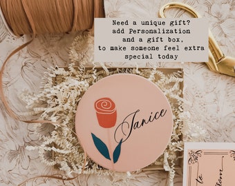 May include: A pink round gift tag with a stylized red rose and the name "Janice" written in cursive. The tag is surrounded by shredded paper in a gift box.