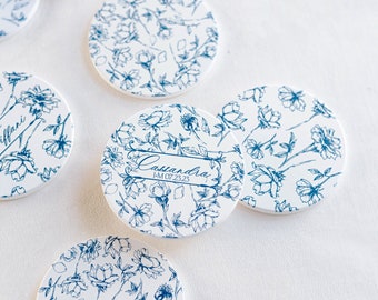 May include: Round white coasters with a blue floral pattern. Some coasters have a name and date written on them.