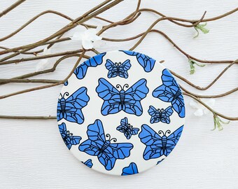 May include: A white round coaster with a blue butterfly pattern. The butterflies are outlined in black and have a white background.