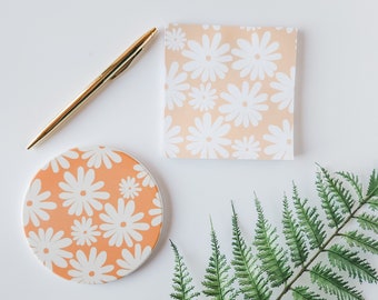 Könnte beinhalten: Ein weißer Hintergrund mit einem goldenen Stift, einem runden orangen Untersetzer mit weißem Gänseblümchen-Druck, einem quadratischen, pfirsichfarbenen Haftnotizblock mit weißem Gänseblümchen-Druck und einem grünen Farn.