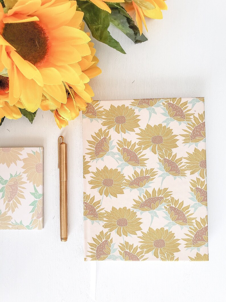 May include: A pink and white notebook with a sunflower pattern. The notebook is closed and lying flat on a white surface. A gold pen is lying next to the notebook. A bouquet of yellow sunflowers is in the top left corner of the image.