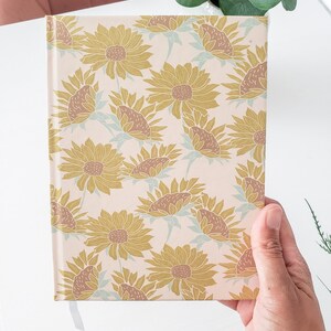 May include: A cream-colored journal with a gold and brown sunflower pattern. The journal has a ribbon bookmark.