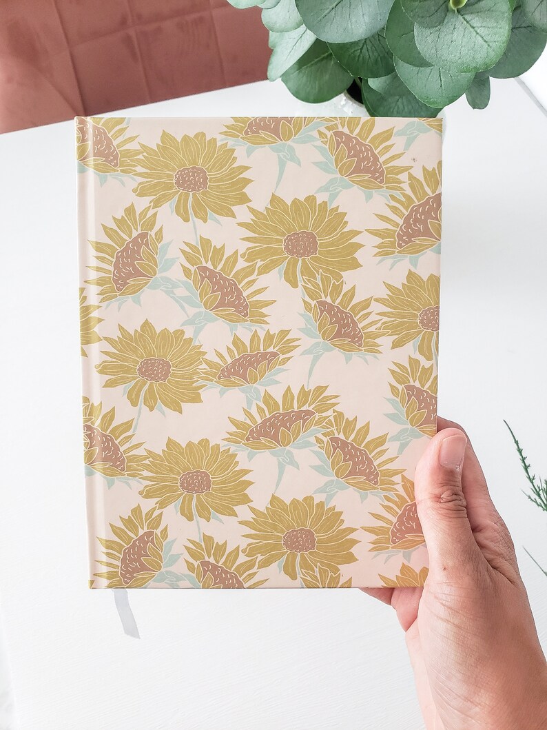 May include: A cream-colored journal with a gold and brown sunflower pattern. The journal has a ribbon bookmark.
