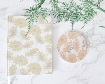 May include: A white journal with a floral pattern of yellow and brown sunflowers. A matching coaster with the same sunflower pattern is also pictured.