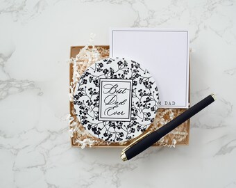 Könnte beinhalten: Ein runder weißer Keramik-Untersetzer mit schwarzem Blumenmuster und dem Text "Best Dad Ever". Der Untersetzer befindet sich in einer braunen Schachtel mit weißem zerknülltem Papier. Ein schwarzer Stift und eine weiße Karte mit dem Text "From Dad" sind ebenfalls in der Schachtel.
