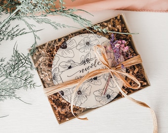 May include: A white ceramic coaster with a floral design and the text "Nicole" in black calligraphy. The coaster is in a clear box with brown shredded paper and a brown ribbon.