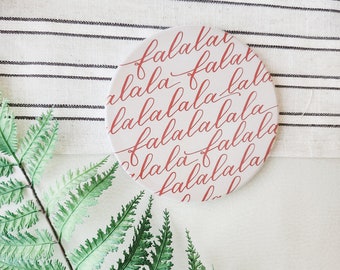 May include: A white round coaster with the words "fa la la" repeated in red cursive writing.