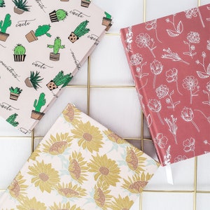 May include: Three different notebooks with patterned covers. The first notebook has a pink cover with a repeating pattern of green cacti and the Spanish word "cactus". The second notebook has a pink cover with a repeating pattern of gold sunflowers. The third notebook has a red cover with a repeating pattern of white flowers.