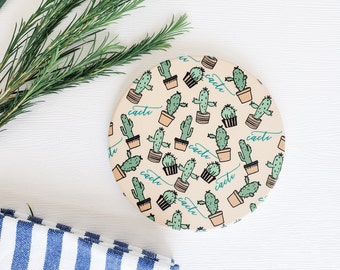 May include: A round, cream-colored coaster with a repeating pattern of green cacti in pots. The word "cacti" is written in a cursive font on the coaster.