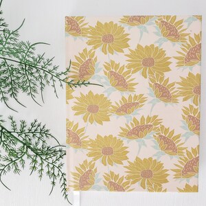 May include: A notebook with a sunflower pattern on a light pink background. The sunflowers are outlined in a darker yellow and have brown centers. The notebook is closed and has a white spine.