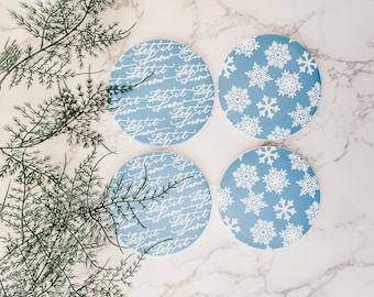 Könnte beinhalten: Vier blaue und weiße Untersetzer aus Keramik. Zwei Untersetzer haben ein Schneeflockenmuster, und zwei Untersetzer haben den Text "Let it snow" in kursiver Schrift wiederholt.