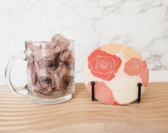 May include: A white ceramic mug with a black line drawing of roses. The mug is filled with crumpled tissue paper. A coaster with a pink and orange floral design is on a wooden surface.