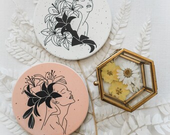 May include: Two round ceramic coasters with a black and white line drawing of a woman's face with flowers in her hair. The coasters are on a white surface with a fern leaf.