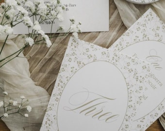 Könnte beinhalten: Elegante Hochzeitspapeterie mit floralem Design. Das Bild zeigt eine Menükarte, Tischkarten mit dem Wort "Three" und einen personalisierten Deckel. Sanfte Farben und zarte Details schaffen eine romantische Ästhetik.