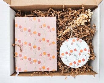 Könnte beinhalten: Eine Geschenkbox gefüllt mit braunem Packpapier, einem rosa Notizbuch mit Blumenmuster, einem weißen Untersetzer mit Blumenmuster und einem goldenen Stift.