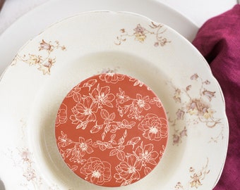Könnte beinhalten: Ein runder, rostfarbener Teller mit weißem Blumendesign liegt auf einem Teller im Vintage-Stil mit floralen Akzenten. Die Teller stehen auf einer weißen Oberfläche, mit einer dunkelbraunen Gabel und einer weinroten Serviette.