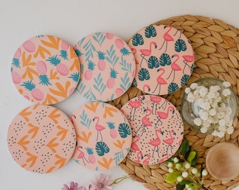 Könnte beinhalten: Sechs runde Untersetzer mit verschiedenen tropischen Designs. Die Untersetzer zeigen rosa Flamingos, Ananas und abstrakte Muster auf einem hellrosa Hintergrund.
