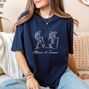 May include: A navy blue t-shirt with a white outline of two people hiking with backpacks and walking sticks. The text "Utreia et Suseia" is printed below the image.