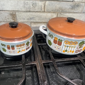 May include: Two white enameled metal pots with matching burnt orange lids. The pots feature a repeating graphic pattern of kitchen utensils, flowers, and spice jars in shades of orange, green, and brown. Each pot has a black knob on the lid.