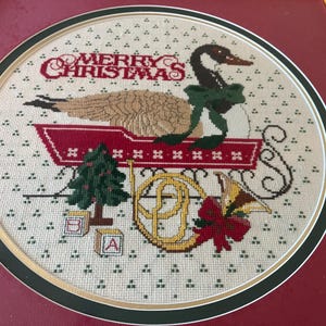 May include: A framed cross-stitch depicting a goose in a sleigh, with a green bow, and the words "MERRY CHRISTMAS" in red. Other elements include a Christmas tree, a horn, and alphabet blocks.