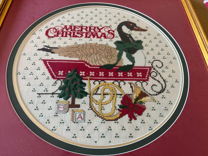 May include: A framed cross-stitch depicting a goose in a sleigh, with a green bow, and the words "MERRY CHRISTMAS" in red. Other elements include a Christmas tree, a horn, and alphabet blocks.