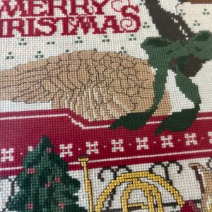 May include: A close-up of a cross-stitch Christmas design. The words "MERRY CHRISTMAS" are stitched in red. A tan bird with a green bow sits atop a red wagon. A green Christmas tree and a gold horn are also visible.