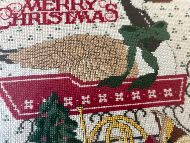 May include: A close-up of a cross-stitch Christmas design. The words "MERRY CHRISTMAS" are stitched in red. A tan bird with a green bow sits atop a red wagon. A green Christmas tree and a gold horn are also visible.