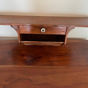 May include: A wall-mounted wooden shelf featuring a dark brown finish, a single horizontal drawer with a small white circular knob, and an open storage compartment located directly beneath the drawer, showcasing a smooth wood grain texture.