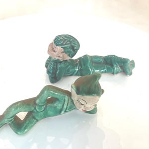 May include: Two ceramic figurines in a reclining pose, each wearing a green suit. One figurine has a pointed hat and painted facial features, while the other has a textured green cap and a neutral face. Both are finished in a glossy green glaze.