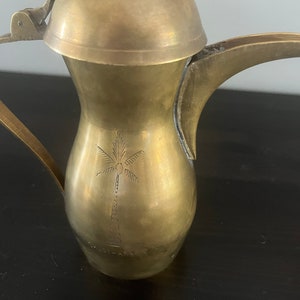 May include: A brass coffee pot with a hinged lid and a palm tree design. The pot has a curved spout and a handle.