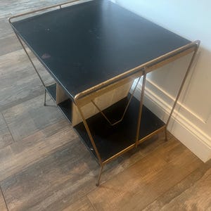 May include: A two-tier side table featuring a rectangular black top and a matching black lower shelf. The frame is constructed from thin, metallic gold-toned metal with angled supports and a raised railing along the top edge of the table.
