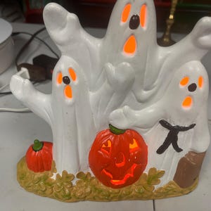 May include: A white ceramic tabletop decoration featuring three ghosts with glowing orange cutouts for eyes and mouths. The ghosts are positioned behind a central orange jack-o'-lantern and a smaller orange pumpkin, all set on a textured base.