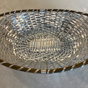 May include: Oval-shaped basket with a woven design. The basket features a silver-colored interior and a brown and silver-colored trim around the rim. The basket is made of a woven material.