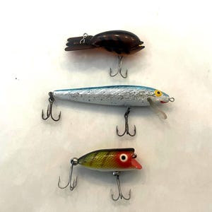 May include: Three fishing lures are displayed against a white background. The lures are in various colors, including brown, silver, blue, red, yellow, and green. Each lure has multiple treble hooks attached.