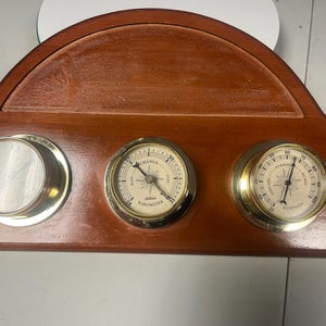 May include: A wooden weather station with three brass-rimmed gauges. The gauges include a barometer, a thermometer, and a hygrometer. The wooden base is a dark brown color.