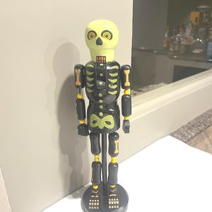 May include: A wooden skeleton figurine with a yellow head and black body. The skeleton has yellow and black accents, including rib details and joint markings. The figurine stands on a black circular base.