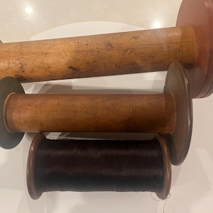 May include: Four antique wooden spools of varying sizes. The largest spool is a light brown color with a dark brown end. The second spool is a medium brown color. The third spool is a dark brown color. The smallest spool is a dark brown color with dark thread.