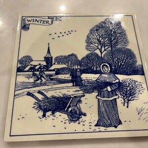 May include: A blue and white ceramic tile with a winter scene. The tile depicts a small village with a church, a woman carrying a bundle of sticks, and a wheelbarrow. The text "WINTER" is written on a banner at the top of the tile.