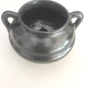 May include: A small, round, black ceramic pot with a glossy finish and two curved handles on opposite sides. The vessel features a wide, open mouth and a smooth, rounded body, showcasing a uniform dark color throughout its entire exterior surface.