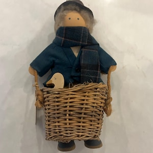 May include: A handmade doll wearing a blue jacket, a plaid scarf, and a black hat. The doll is holding a wooden duck and sitting in a wicker basket.