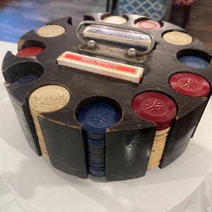 May include: A vintage wooden poker chip carousel with a dark finish. It holds stacks of red, blue, and cream-colored poker chips. The carousel has a metal handle and slots for chip storage, along with two boxes of playing cards.