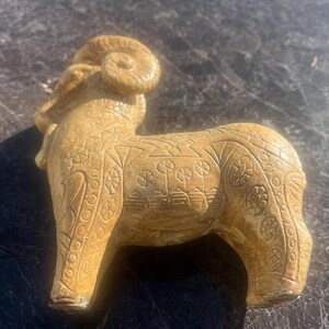 May include: A tan-colored ram figurine with detailed carvings. The sculpture features curled horns and intricate patterns on its body, including floral and geometric designs. The ram is standing on all fours, showcasing a detailed, textured surface.