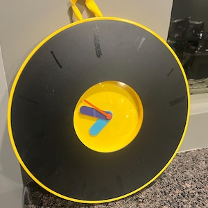 May include: A round, modern wall clock with a black face and yellow trim. The clock has a yellow center with blue, orange, and purple hands. The clock is hanging by a yellow strap.