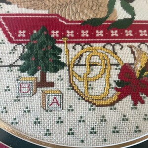 May include: Cross-stitch embroidery with a Christmas theme. The design includes a green Christmas tree, a golden French horn, a red bow, and alphabet blocks. The background is a light beige fabric with small green cross-stitch details.
