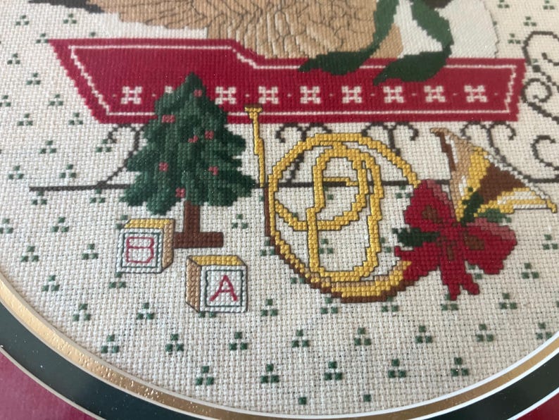 May include: Cross-stitch embroidery with a Christmas theme. The design includes a green Christmas tree, a golden French horn, a red bow, and alphabet blocks. The background is a light beige fabric with small green cross-stitch details.