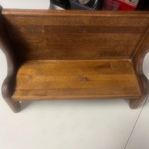 May include: A small, wooden bench with a curved back and armrests. The bench is made of a light brown wood with a visible wood grain. The bench has a rectangular seat and is supported by four short legs. The overall design is simple and rustic.